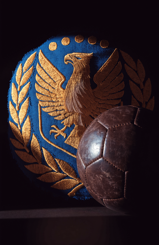 Italy's deep azure sits on rich embroidered fabric beside a worn leather ball, lit from one side